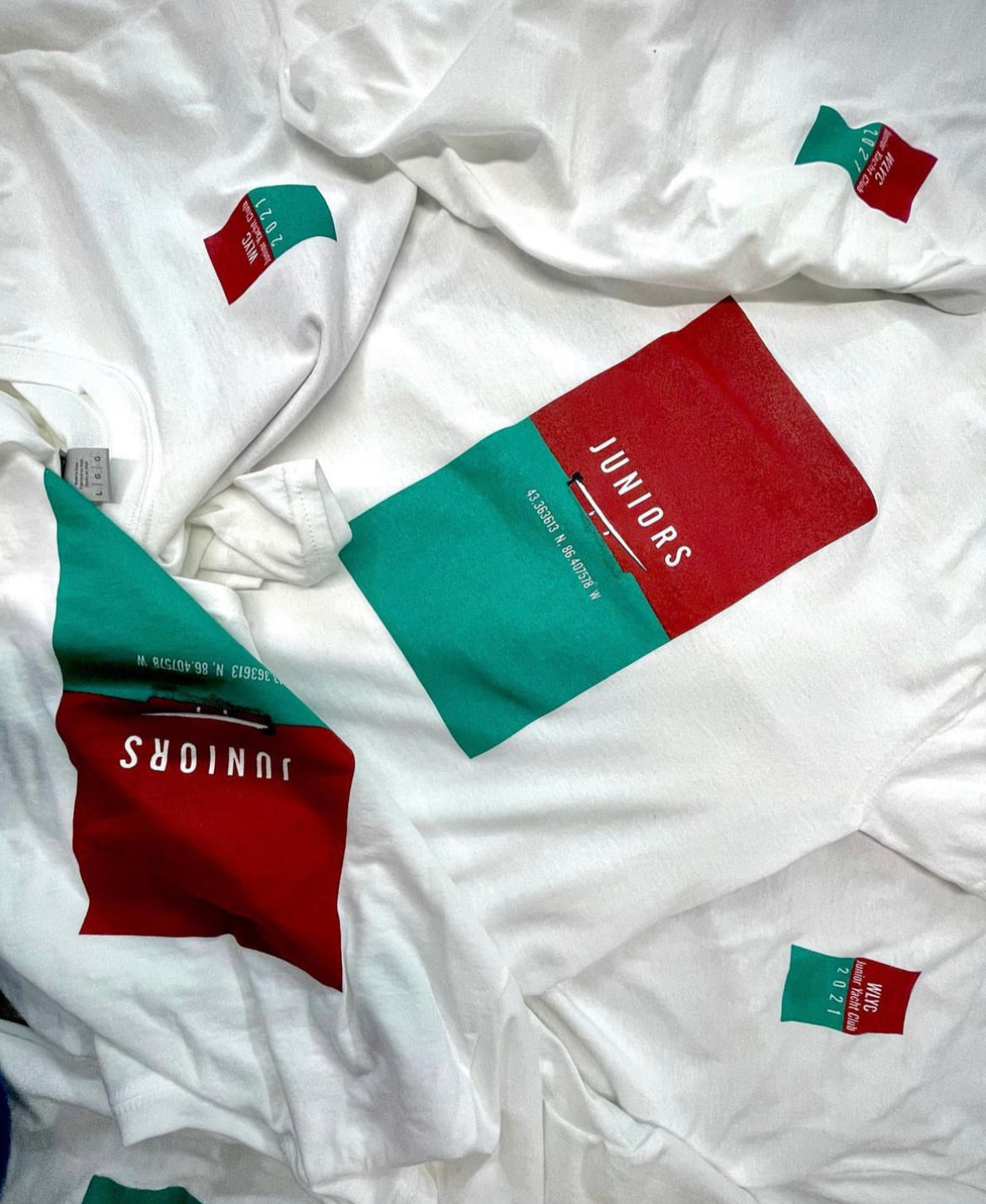 Pile of Custom Screen Printed Tee Shirts for Juniors - Red and Green Design With Red Boat on Green Lake. Custom Shirts for Michigan Locals